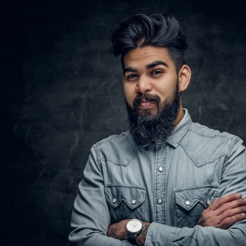 Studio portrait of stylish bearded Indian man in a blue shirt over grey background.